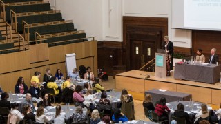 An auditorium packed with conference delegates seated at round tables. A speaker delivers a speech from a podium on a stage.